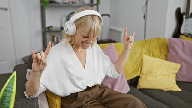 Blonde woman enjoying music at home, showing a rock gesture, wearing white headphones and sitting on a grey sofa with colorful pillows in a cozy living room