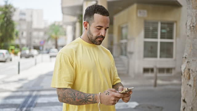 Handsome hispanic man with beard and tattoos using smartphone on a sunny urban street.