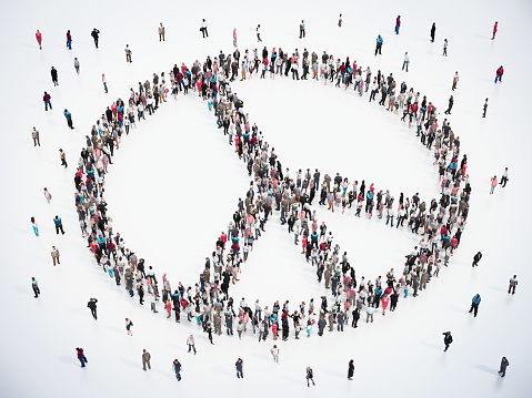 A large group of individuals stands in formation, creating the distinct outline of a peace symbol. The stark white background enhances the visibility of the human pattern, showcasing a symbolic gesture of unity and non-violence.