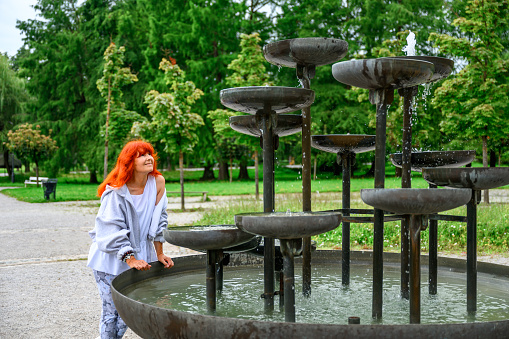 Senior woman with long red dyed hair standing by the water fountain in a public park.
