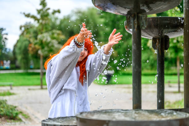 Cheerful senior woman with long red dyed hair splashing water at the water fountain in a public park.
