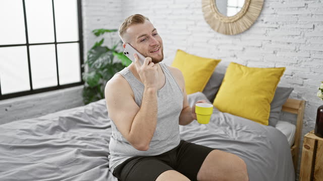A smiling young man with a beard enjoys a morning phone call while holding a cup in a stylish bedroom.
