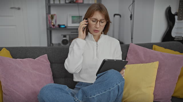 A young woman multitasks in her modern living room, talking on the phone and using a tablet.