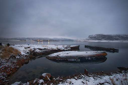 Sunken boats lie in a serene, snow draped bay as dusk falls on the quiet landscape