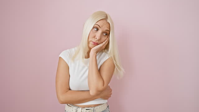 Depressed and unhappy, young blonde woman, arms crossed, tired and bored, lost in troublesome thoughts, isolated over a pink background