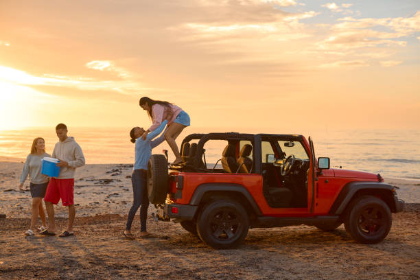 Young Couple With Friends Unloading Cool Box From Car At Beach At Sunrise