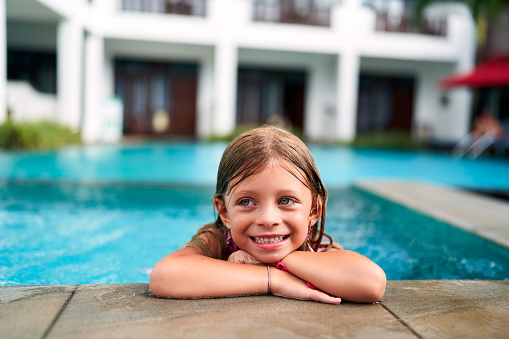 Child enjoys summer water fun, swim in kid-friendly pool. Smiling little girl rests on pool edge at sunny resort. Vacation leisure, happy childhood, family travel at tropical hotel. Aquatic play.
