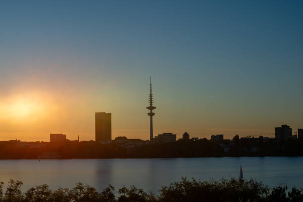 The golden hues of the setting sun wash over the cityscape, casting a serene glow on the skyline dominated by a towering silhouette. The calm waters provide a mirror-like reflection, interrupted only by the gentle passage of a sailboat. It's an image that captures the peaceful coexistence of urban architecture and nature's quiet end-of-day beauty