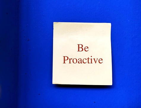 Note stick on blue wall with text written BE PROACTIVE , different traits - one focuses on eliminating problems before happened and one approach is based on responding to events after they have