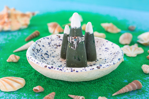 Handmade silver ring with blue stone on a ceramic ring holder and artificial green sand with seashells on a blue background.