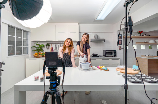 Two asian women in an apron, smiling and cooking in a kitchen with camera and lighting equipment for video and photo shoot, recording vlog video for social blogger.