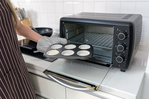 A person in an apron and oven gloves, placing a muffin tray with batter into a black toaster oven in a white kitchen.