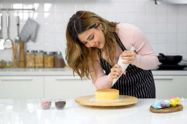 istock Young woman in an apron using a piping bag, pipe the icing onto the cake, creating a decorative pattern on the top of a round, light-colored cake that's placed on a wooden turntable or cake stand. 2132406743