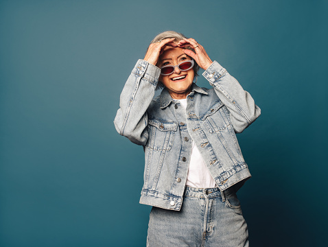 Happy, mature woman with grey hair stands confidently in a studio, wearing a fashionable denim outfit and eyeglasses. Her playful smile and carefree demeanor capture the essence of a carefree and fun-loving older individual.
