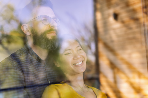 Mid adult interracial couple standing by the window, looking outside. It's a beautiful sunny day, they look relaxed and happy. The photo is taken through the window.