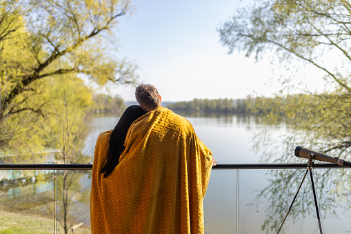Mid adult interracial couple enjoying their time on a balcony of the house by the lake. They are wrapped in a blanket, it's a beautiful sunny day, they look relaxed, happy and in love.