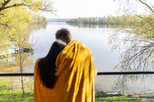 Mid adult interracial couple enjoying their time on a balcony of the house by the lake. They are wrapped in a blanket, it's a beautiful sunny day, they look relaxed, happy and in love. Focus is on background