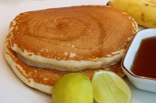 Stock photo showing close-up view of American-style, fluffy pancakes stack on white plate with split ramekin of honey and chocolate spread, lemon wedges and a banana.