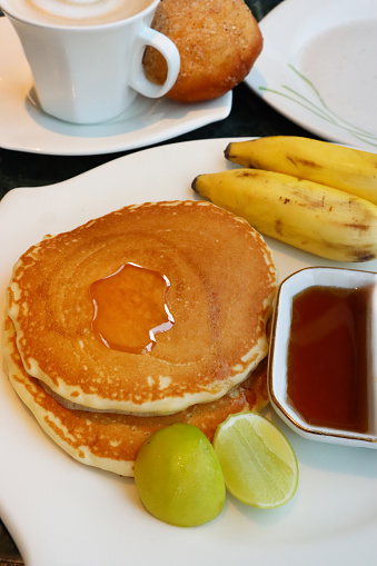 Stock photo showing close-up, elevated view of American-style, fluffy pancakes stack on white plate with split ramekin of honey and chocolate spread, lemon wedges and bananas.