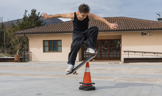 A skateboarder in a tank top jumps over a traffic cone, showcasing precision and control in an urban setting.
