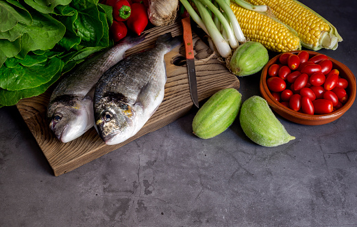 Fish sea breams(Sparus aurata) on a chopping board, knife and different vegetables on a dark background close-up