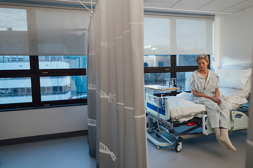 A medium wide angle view of a woman sitting on the edge of her hospital bed before she goes for some treatment for her breast cancer. She has a contemplative look on her face as she anticipates the treatment she is about to receive.\n\nVideos are available for this scenario