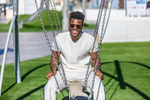 An African American man in his 30s sitting on a swing in a public park, surrounded by green grass.