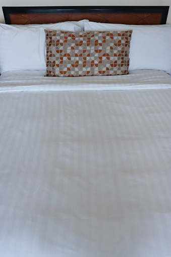 Stock photo showing close-up view of beige, orange and white patterned cushion leaning against a pile of white hotel pillows resting on wooden headboard on hotel room duvet bedding.