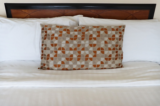 Stock photo showing close-up view of beige, orange and white patterned cushion leaning against a pile of white hotel pillows resting on wooden headboard on hotel room duvet bedding.