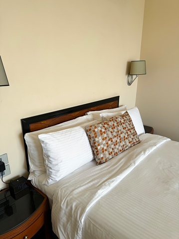 Stock photo showing close-up view of beige, orange and white patterned cushion leaning against a pile of white hotel pillows resting on wooden headboard on hotel room duvet bedding. In the bedroom beside the bed stands a wooden nightstand with drinking glasses turned upside down and an electric wall lamp.