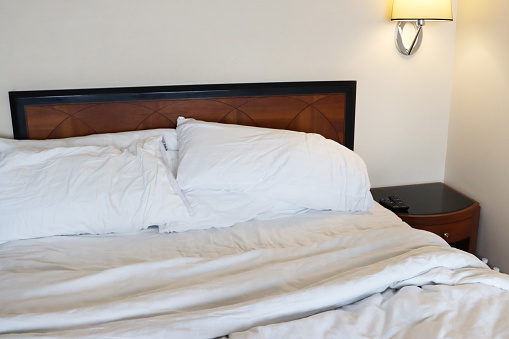 Stock photo showing close-up view of remade double bed with crumpled, white bedding after being slept in. In the bedroom beside the bed stands a wooden nightstand and electric wall lamp.