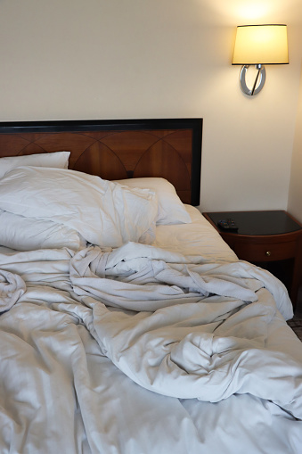 Stock photo showing close-up view of unmade double bed with crumpled, white bedding. In the bedroom beside the bed stands a wooden nightstand and electric wall lamp.