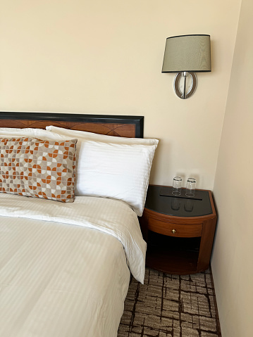 Stock photo showing close-up view of beige, orange and white patterned cushion leaning against a pile of white hotel pillows resting on wooden headboard on hotel room duvet bedding. In the bedroom beside the bed stands a wooden nightstand with drinking glasses turned upside down and an electric wall lamp.