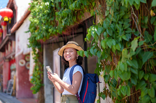 A solo Asian woman traveler, with a backpack and straw hat, uses her smartphone while exploring Georgetown, Penang. She travels light, relying on technology and staying connected during her solo trip.