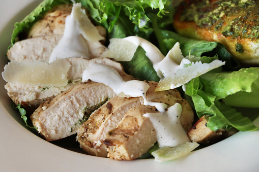 Stock photo showing close-up, elevated view of healthy chicken Caesar salad served at a restaurant, shaved parmesan cheese, fried chicken slices, crispy green romaine lettuce leaves, toasted garlic bread slices spread with green pesto and salad dressing.