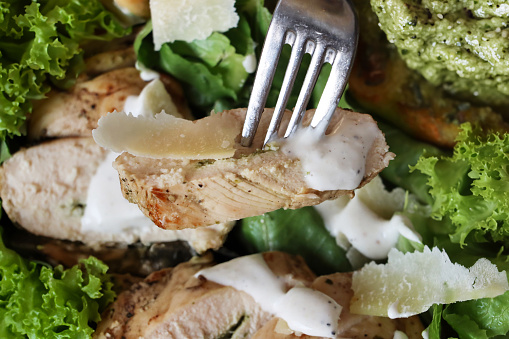 Stock photo showing close-up, elevated view of healthy chicken Caesar salad served at a restaurant, shaved parmesan cheese, fried chicken slices, crispy green romaine lettuce leaves, toasted garlic bread slices spread with green pesto and salad dressing.