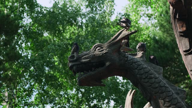 Pigeons sitting on a dragon statue in the city park. Urban birds hide from the summer heat in the shade of green foliage.