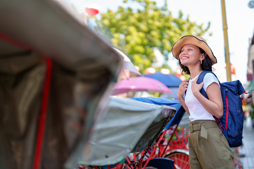 A cheerful solo Asian woman traveler, wearing a stylish straw hat adorned with a colorful ribbon and sporting a comfortable backpack, is seen joyfully exploring the vibrant streets of Georgetown, Penang.