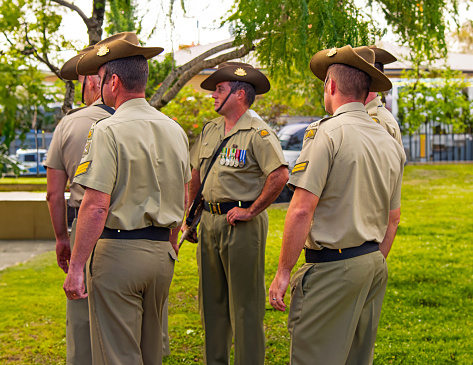 Candid photograph of a group of five male Australian Army soldiers from the Royal Australian Corps of Engineers (RAE) wearing ceremonial uniform, defence service medals, iconic slouch hat, and F88 Austeyr rifles awaiting their turn to march onto the Cenotaph memorial at an ANZAC Day, Remembrance Day or Veteran's Day parade service in Australia.