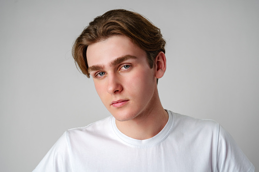 A young man wearing a white t-shirt is gazing directly at the camera, his expression neutral and focused. He stands in a simple setting, with no other discernible objects or people around him.