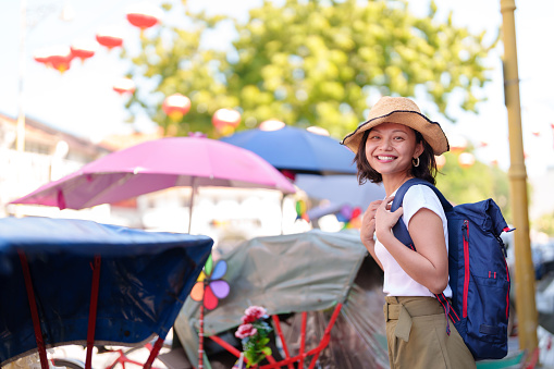 A cheerful solo Asian woman traveler, wearing a stylish straw hat adorned with a colorful ribbon and sporting a comfortable backpack, is seen joyfully exploring the vibrant streets of Georgetown, Penang.