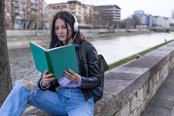 istock Young beautiful female university student with backpack is enjoying near the river. She is wearing headphones and leather jacket while waiting for her college class to start. 2132847201