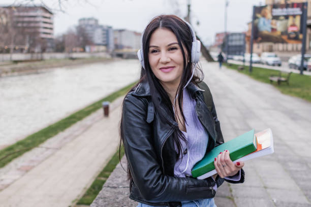 istock Young beautiful female university student with backpack is enjoying near the river. She is wearing headphones and leather jacket while waiting for her college class to start. 2132851891