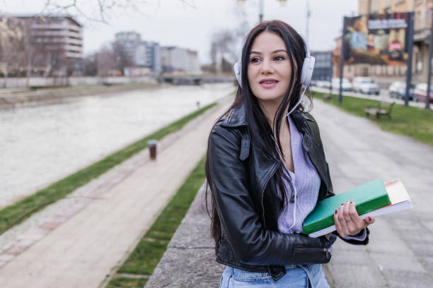 istock Young beautiful female university student with backpack is enjoying near the river. She is wearing headphones and leather jacket while waiting for her college class to start. 2132853469