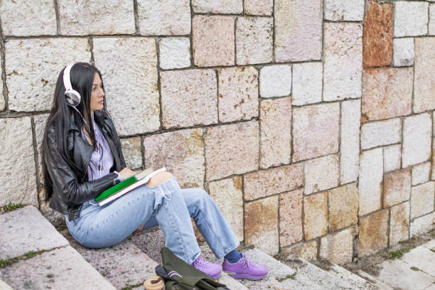 istock Young beautiful female university student with backpack sitting on stone steps. She is wearing headphones and leather jacket while waiting for her college class to start. 2132856081