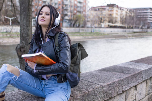 istock Young beautiful female university student with backpack is enjoying near the river. She is wearing headphones and leather jacket while waiting for her college class to start. 2132860197