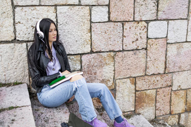 istock Young beautiful female university student with backpack sitting on stone steps. She is wearing headphones and leather jacket while waiting for her college class to start. 2132864304