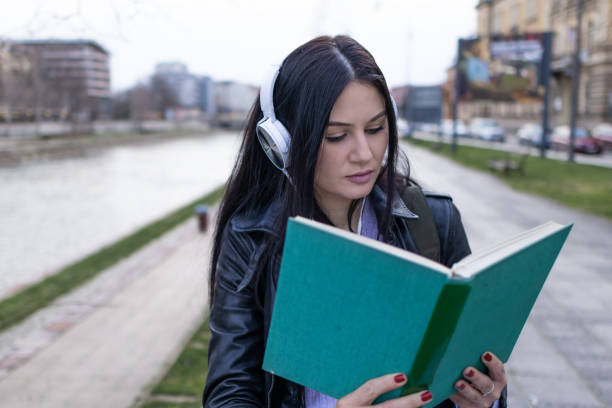 istock Young beautiful female university student with backpack is enjoying near the river. She is wearing headphones and leather jacket while waiting for her college class to start. 2132870110