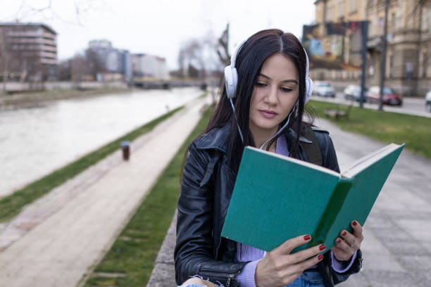 istock Young beautiful female university student with backpack is enjoying near the river. She is wearing headphones and leather jacket while waiting for her college class to start. 2132871144