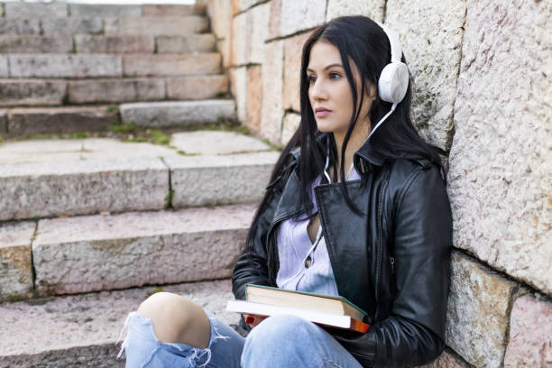 istock Young beautiful female university student with backpack sitting on stone steps. She is wearing headphones and leather jacket while waiting for her college class to start. 2132874278